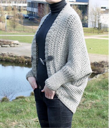 Person trägt hellgraue gehäkelte Oversize-Jacke mit Knebelverschluss vor einem See.