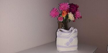 Cremeweiße gehäkelte Tasche mit lila Streifen und Schleife auf heller Ablage neben Vase mit bunten Blumen.