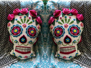 Zwei gehäkelte weiße Totenkopf-Kissen mit pinken Rosen, blauen Blumenaugen und sichtbaren Zähnen auf gemusterten Decken