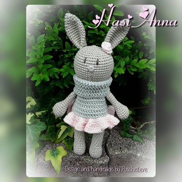 Graues Amigurumi-Häschen mit rosa Rüschenrock und Schal, vor grünen Blättern