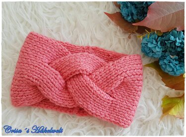 Gestricktes korallenfarbenes Stirnband mit Zopfmuster auf weißem Fellimitat, daneben blaue Blüten und Herbstlaub