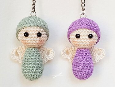 Zwei gehäkelte Amigurumi-Engelchen als Schlüsselanhänger in Mint und Flieder mit Flügeln, Sicherheitsaugen und Metallkette auf hellem Hintergrund.