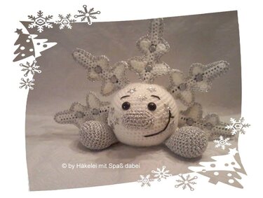 Weißes gehäkeltes Amigurumi mit lachendem Gesicht, silberumrandeten Schneeflocken-Elementen und neutralem Hintergrundrahmen