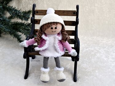 Gehäkelte Amigurumi-Puppe mit braunen Locken, rosa Jacke, weißer Mütze und Stiefeln auf einer Miniaturbank