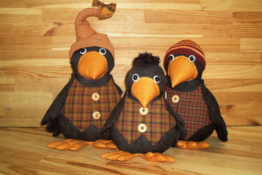 Drei genähte Krähenfiguren mit karierten Westen und orangefarbenen Schnäbeln vor Holzhintergrund.