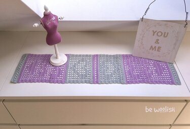 Gehäkelter Tischläufer in Flieder und Grau auf weißer Kommode, dekoriert mit einer Schneiderpuppe und einem Holzschild 'YOU & ME'.