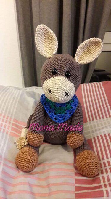 Gehäkelter Amigurumi-Esel in Braun und Beige mit blau-grünem Halstuch auf gestreifter Decke