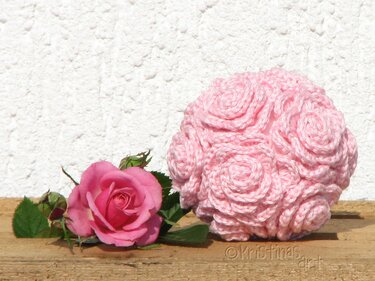 Rosa gehäkelte Rosenkugel neben einer echten rosa Rose auf Holz