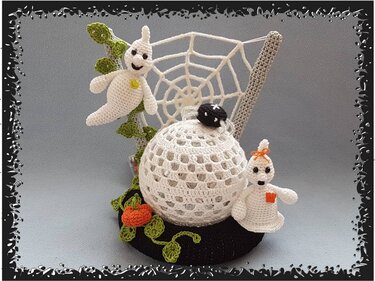 Gehäkelte Halloween-Dekoration mit zwei weißen Gespenstern, Spinnennetz, gehäkelter Kugel mit schwarzer Spinne, Kürbis und grünen Blättern auf schwarzer Basis.