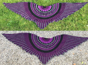 Wide wing-shaped crocheted shawl in purple, black, and grey with scalloped textured edges, laid flat on grass and gravel.