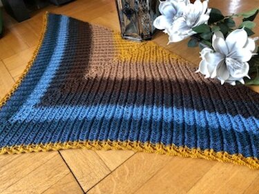 Gehäkeltes gestreiftes Dreieckstuch in Blau, Braun und Senfgelb auf Holzfußboden neben weißen Blumen.