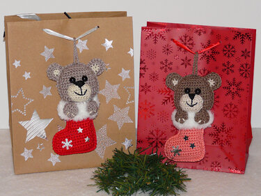 Zwei gehäkelte Teddybären in roten und rosa Weihnachtsstrümpfen an Geschenkbeuteln mit Stern- und Schneeflockenmuster, Tannengrün im Vordergrund