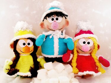Three crocheted amigurumi dolls in winter hats and coats with white cubes on a snowy surface.