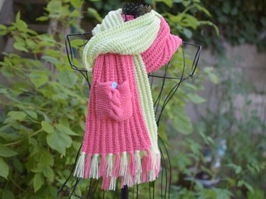 Gestrickter pink-hellgrüner Schal mit aufgesetzter Tasche und Fransen auf einer Drahtpuppe vor grünem Hintergrund.