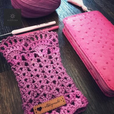 Pinke gehäkelte Armstulpen mit Glitzergarn, Häkelnadeln und Etui auf dunklem Holzhintergrund.
