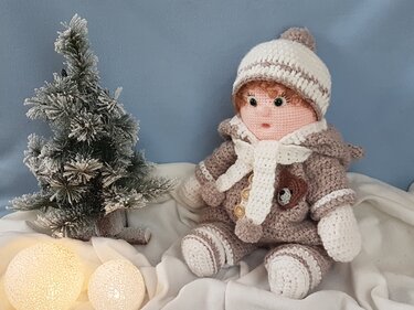 Gehäkelte Amigurumi-Puppe in Winterkleidung mit Mütze und Schal sitzt neben einem kleinen, beschneiten Tannenbaum und leuchtenden Kugeln auf einer weißen Decke