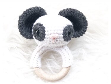 Gehäkelte Panda-Rassel mit schwarzen Ohren und Augen sowie rosa Nase, befestigt an einem hellen Holzring auf weißem Fell.