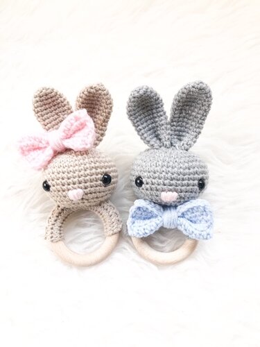 Zwei gehäkelte Hasen-Beißringe mit Holzringen, links beige mit rosa Schleife, rechts grau mit blauer Schleife auf hellem Hintergrund