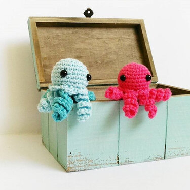 Zwei gehäkelte Amigurumi-Kraken (hellblau und pink) auf einer offenen Holzkiste.