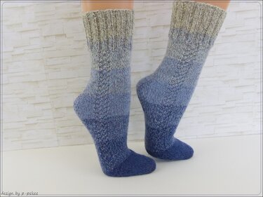 Zwei F
ße tragen handgestrickte Socken mit sanftem Farbverlauf von Beige zu Blau vor heller Wand