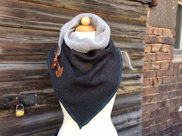 Dunkelgraues Dreieckstuch mit hellgrauem Fellfutter, türkiser Kettelnaht und lederähnlichen Verschlüssen auf Schaufensterpuppe vor Holzfassade