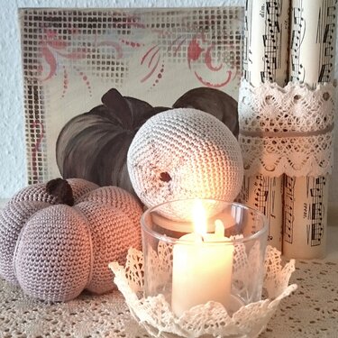 Zwei gehäkelte Kürbisse in Pastellfarben neben brennender Kerze im Glas und Spitzenunterlage, im Hintergrund gerollte Notenseiten und Gemälde
