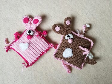 Zwei gehäkelte Schnuffeltücher in Rosa und Braun mit Hasenohren, flauschigem Bauchfleck und Augen auf hellem Untergrund