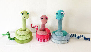 Drei gehäkelte Amigurumi‑Schlangen (grün, pink, blau) aufgerollt auf weißer Fläche