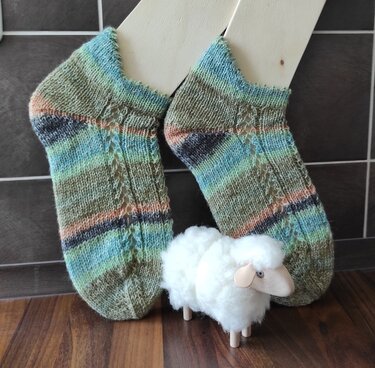 Paar gestrickter knöchelanger Socken in grünen, blauen und pfirsichfarbenen Streifen mit seitlichem Ajourmuster auf Holz-Sockenspannern, davor kleine Schäfchenfigur
