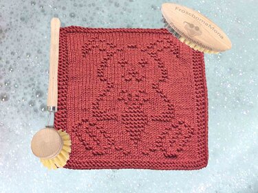 Quadratisches gestricktes Deckchen in Dunkelrot mit reliefartigem Osterhasen-Motiv, daneben Holzbürsten