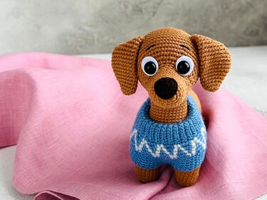 Brown crocheted dachshund wearing a blue sweater with white zigzags on pink fabric.
