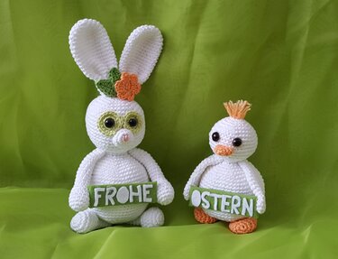 Zwei weiße gehäkelte Amigurumi-Figuren, Hase mit langen Ohren und Küken, halten grüne Schilder 'FROHE OSTERN' vor grünem Hintergrund