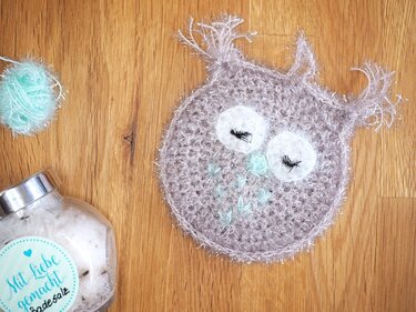 Gehäkelte Eule aus flauschigem grauem Garn mit weißen Augen und mintgrünen Details auf Holz