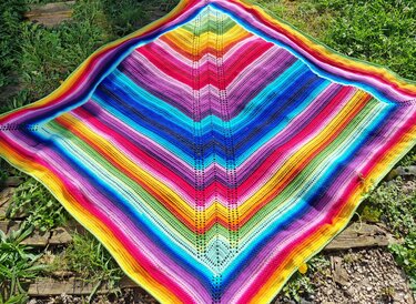 Gehäkelte Decke mit Zickzackmuster in Regenbogenfarben auf Gras und Holzplanken