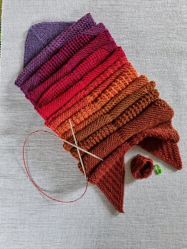 Halbfertiger gestrickter Schal mit Farbverlauf von Lila über Rot bis Orange-Braun, Texturstreifen, Rundstricknadeln und Garnknäuel auf hellem Untergrund