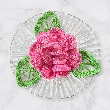 Gehäkelte pinke Rose mit drei grünen Blättern auf einem runden Metallring mit weißem Wickelgarn