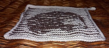 Gestrickter braun-weißer Topflappen mit Wellenmuster und Aufhängeschlaufe auf Weidengeflecht