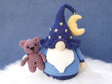 Gehäkelter Wichtel mit blauem Sternenhut und kleinem braunem Teddybär auf weißem Fell vor blauem Hintergrund.