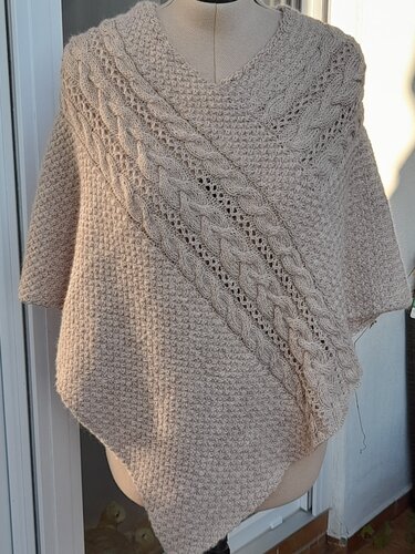 Gestrickter hellbeiger Poncho mit V-Ausschnitt, diagonalen Zopf- und Ajourmustern auf einer Schneiderpuppe.