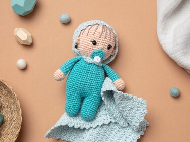 Gehäkelte Amigurumi-Babypuppe in türkis mit Schnuller und Häkeltuch auf beigem Hintergrund