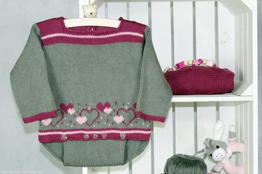 Gestrickter Babystrampler in Graugrün mit rosa Herzbordüre auf einem Regal