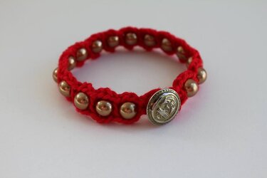Rotes gehäkeltes Armband mit goldfarbenen Perlen und ovalem Knopf mit Anker‑Motiv auf hellem Hintergrund.
