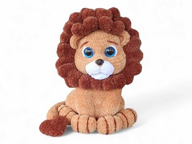 Sitting crocheted lion amigurumi with a fluffy brown mane and blue eyes on a white background.