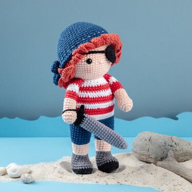 Gehäkelte Amigurumi-Piratenpuppe mit Augenklappe und Schwert auf Sand vor blauem Hintergrund.