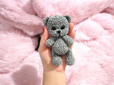 Kleiner grauer gehäkelter Teddybär in einer Hand vor verschwommenem rosafarbenem Plüschhintergrund.