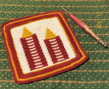 Häkelquadrat mit zwei stilisierten Kerzen in Rot und Gelb auf grün gemustertem Stoff mit Häkelnadel