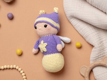 Von oben fotografiertes Amigurumi-Püppchen mit Stern-Applikation und Flügeln in Lila und Gelb, umgeben von Holzperlen und Beißring