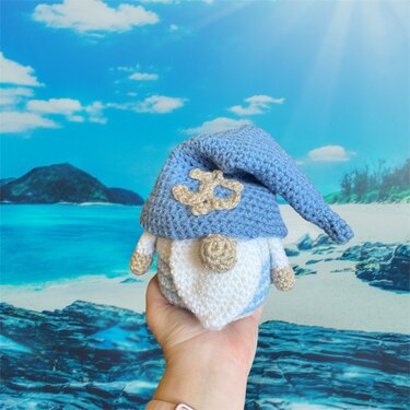 Gehäkelter Wichtel mit blauer Mütze und Anker in einer Hand vor Strandkulisse.