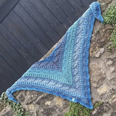 Triangular crochet shawl in blue and teal gradient with intricate lace and textured stitches, draped over a stone wall.