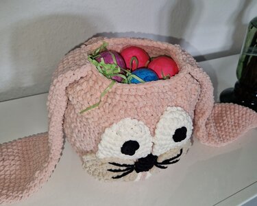 Gehäkelter rosa Hasenkorb mit weißen Augen, schwarzer Nase und Schnurrhaaren, gefüllt mit bunten Ostereiern und grünem Ostergras.